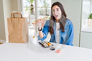 Beautiful young woman eating asian sushi from home delivery scared in shock with a surprise face, afraid and excited with fear