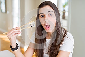 Beautiful young woman eating asian sushi using chopsticks scared in shock with a surprise face, afraid and excited with fear