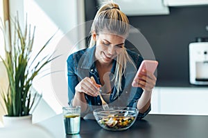 Beautiful young woman eating a bowl of salad while using her mobile phone in the kitchen at home