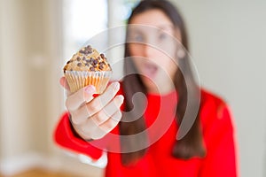 Beautiful young woman eating chocolate chips muffin scared in shock with a surprise face, afraid and excited with fear expression