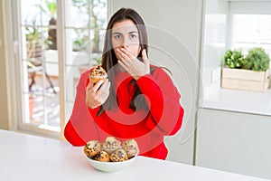 Beautiful young woman eating chocolate chips muffins cover mouth with hand shocked with shame for mistake, expression of fear,