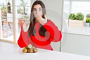 Beautiful young woman eating half fresh green kiwi cover mouth with hand shocked with shame for mistake, expression of fear,