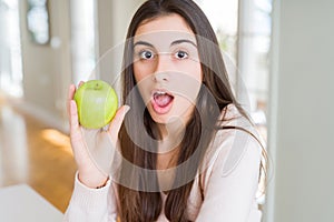 Beautiful young woman eating healthy green apple fruit scared in shock with a surprise face, afraid and excited with fear