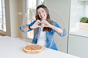 Beautiful young woman eating homemade tasty pizza at the kitchen smiling in love showing heart symbol and shape with hands