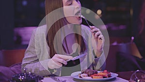 Beautiful young woman eating meat grilled with vegetable in the modern restaurant. The girl cuts one piece of meat and