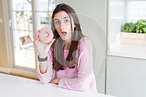 Beautiful young woman eating pink chocolate chips donut scared in shock with a surprise face, afraid and excited with fear