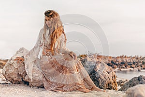 Beautiful young woman in elegant dress sitting on the beach at sunset