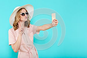 Beautiful young woman in elegant pale pink dress, sunglasses and summer hat taking selfie. Studio portrait of fashionable woman.