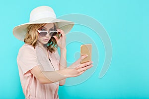 Beautiful young woman in elegant pale pink dress, sunglasses and summer hat taking selfie. Studio portrait of fashionable woman.