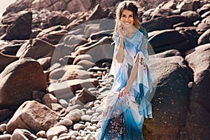 Beautiful young woman in elegant stylish dress on stone beach at sunset