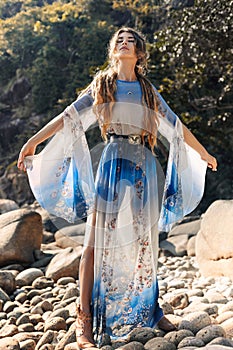 Beautiful young woman in elegant stylish dress on stone beach at sunset