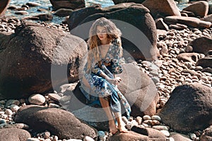 Beautiful young woman in elegant stylish dress on stone beach at sunset