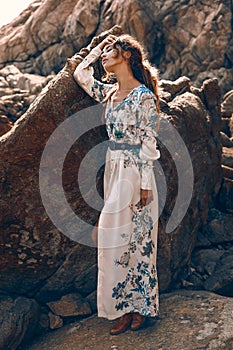 Beautiful young woman in elegant stylish dress on stone beach at sunset