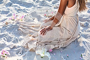 Beautiful young woman in elegant white dress sitting on sand at sunset close up