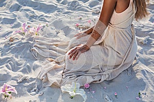 Beautiful young woman in elegant white dress sitting on sand at sunset close up