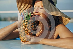 Beautiful young woman enjoying cocktail in pineapple, relaxing in pool on sunny summer vacation. Portrait of girl in hat with