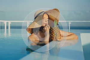 Beautiful young woman enjoying cocktail in pineapple, relaxing in pool on sunny summer vacation. Portrait of girl in hat with