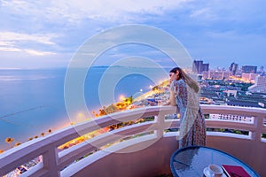 Beautiful young woman enjoying the spectacular nightlife Pattaya city from terrace hotel, with light of twilight, sea and beach in