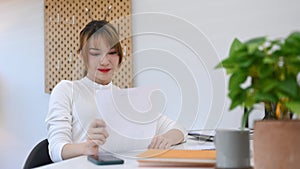 Beautiful young woman entrepreneur in white sweater reading document and using laptop during working from home