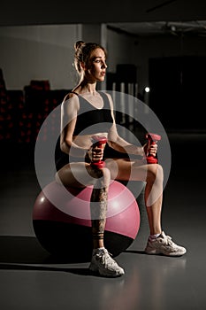 Beautiful young woman exercising with dumbbells on fitness ball at gym against mirror