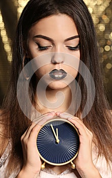 Beautiful young woman with extravagant makeup holding clock against defocused lights