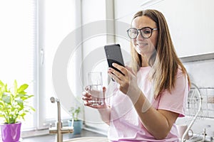 Beautiful young woman in eyeglasses drinking water while using mobile phone at home kitchen. Brunette girl reading or typing a