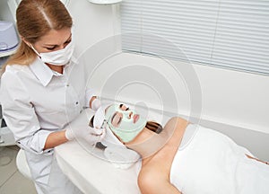 Beautiful young woman with facial mask in beauty salon. Girl getting beauty treatment facial care