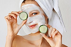 Beautiful young woman with facial mask on her face holding slices of cucumber. Skin care and treatment, spa, natural beauty and