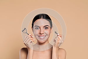 Beautiful young woman with false eyelashes and curler on beige background