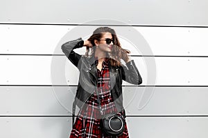 Beautiful young woman in fashion casual red-black youth outfit in stylish sunglasses with leather bag straightens hairstyle near