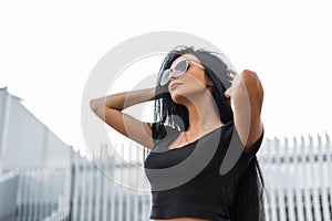 Beautiful young woman fashion model with chic black hair in a trendy black t-shirt in stylish dark sunglasses posing outdoors