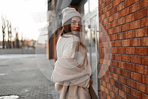 Beautiful young woman in a fashionable gray hat in a stylish knitted sweater in a youth spring eco fur coat posing near a vintage