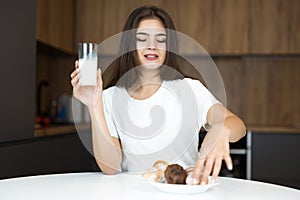 Beautiful young woman feeling hungry eating cupcakes and croissants and drinking milk for breakfast in the kitchen