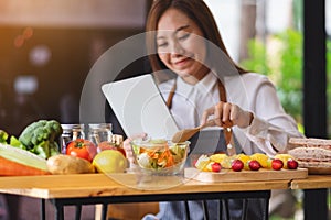 A beautiful young woman following recipe on digital tablet while cooking salad and sandwich in the kitchen, online learning