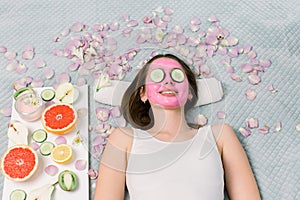 Beautiful young woman is getting facial clay mask at spa, lying with cucumbers on eyes