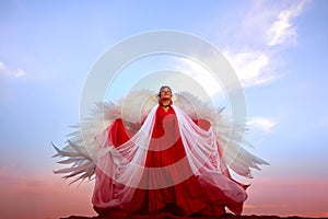 Beautiful young woman or girl with curly hair and in red dress with a light flying fabric and white wings on the sand on