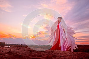 Beautiful young woman or girl with curly hair and in red dress with a light flying fabric and white wings on the sand on