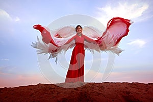 Beautiful young woman or girl with curly hair and in red dress with a light flying fabric and white wings on the sand on