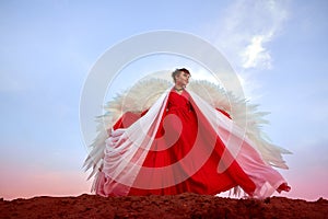 Beautiful young woman or girl with curly hair and in red dress with a light flying fabric and white wings on the sand on