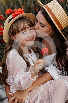 Beautiful young woman with girl in field with poppies, mother and daughter in white dresses and straw hats in evening at sunset,