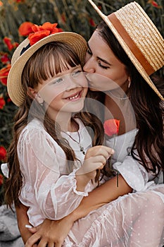 Beautiful young woman with girl in field with poppies, mother and daughter in white dresses and straw hats in evening at sunset,