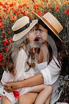 Beautiful young woman with girl in field with poppies, mother and daughter in white dresses and straw hats in evening at sunset,