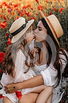 Beautiful young woman with girl in field with poppies, mother and daughter in white dresses and straw hats in evening at sunset,