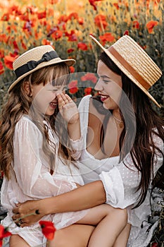 Beautiful young woman with girl in field with poppies, mother and daughter in white dresses and straw hats in evening at sunset,