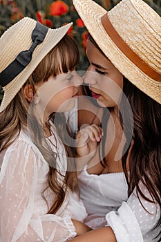 Beautiful young woman with girl in field with poppies, mother and daughter in white dresses and straw hats in evening at sunset,