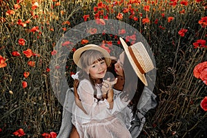 Beautiful young woman with girl in field with poppies, mother and daughter in white dresses and straw hats in evening at sunset,