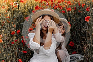 Beautiful young woman with girl in field with poppies, mother and daughter in white dresses and straw hats in evening at sunset,