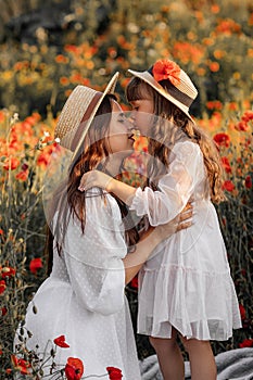 Beautiful young woman with girl in field with poppies, mother and daughter in white dresses and straw hats in evening at sunset,