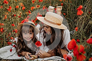 Beautiful young woman with girl in field with poppies, mother and daughter in white dresses and straw hats in evening at sunset,