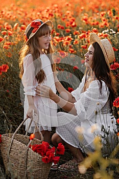 Beautiful young woman with girl in field with poppies, mother and daughter in white dresses and straw hats in evening at sunset,